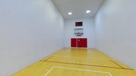 This single-floor indoor sports court offers a versatile athletic environment designed to accommodate multiple sports and activities. The court features a classic hardwood floor finished in a warm golden tone, with clearly marked white and red boundary lines supporting games such as basketball and racquetball. The soaring ceiling height provides ample vertical clearance for athletic play, while recessed square lighting panels illuminate the space evenly, minimizing shadows across the playing surface. A regulation-style basketball hoop with a clear backboard is mounted on the far wall, accompanied by a red padded wall protector beneath it — a safety feature that lines the base of the end wall. The surrounding walls are finished in clean, bright white, contributing to a crisp, well-lit atmosphere that enhances visibility throughout the court. The overall design is functional and straightforward, with an emphasis on maximizing usable play space. Whether used for competitive games or casual recreation, this indoor court delivers a dedicated, purpose-built environment suited for a wide range of athletic activities year-round.