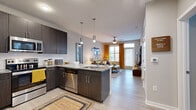 Located at 2120 N Brevard St in Charlotte, NC, this 1-bedroom, 1-bathroom residence offers a thoughtfully designed open-concept floor plan that maximizes its efficient footprint. The modern kitchen is a standout feature, showcasing dark espresso cabinetry, granite countertops, a full suite of stainless steel appliances, subway tile backsplash, and a peninsula with bar seating — all illuminated by contemporary pendant lighting. The kitchen flows seamlessly into the living area, which is bathed in natural light through large windows and a glass door providing outdoor access. Light wood-style flooring runs throughout the main living spaces, enhancing the airy, cohesive feel. The spacious bedroom features plush carpet, generous windows overlooking outdoor views, and ample room for a full furniture arrangement. The bathroom is well-appointed with a granite vanity, dark cabinetry matching the kitchen, a tiled soaking tub and shower combination, and built-in shelving for added storage. A ceiling fan in the living room and consistent modern finishes throughout lend the space a polished, contemporary character suited for comfortable urban living in Charlotte's NoDa neighborhood.