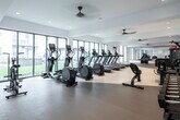 This well-appointed fitness center is designed to support a comprehensive workout experience within a bright, open single-floor layout. The space is defined by its clean, contemporary aesthetic, featuring white walls, smooth flooring in warm neutral tones, and recessed lighting complemented by ceiling fans that maintain airflow throughout.

The cardio zone is prominently featured along one side of the room, housing a generous row of treadmills and elliptical machines, all equipped with individual digital displays. Large floor-to-ceiling windows line the perimeter, inviting abundant natural light and offering views of the surrounding outdoor amenity areas, including landscaped grounds and a pool area visible from multiple vantage points.

On the opposite end, a strength-training section offers a cable and pulley machine, adjustable benches, and a well-stocked dumbbell rack. Large wall-mounted mirrors amplify the sense of space and allow users to monitor their form during workouts. Additional equipment includes air bikes and medicine balls for varied training options.

Glass partitions connect the fitness center to adjacent common areas, maintaining visual openness while defining the workout space. The overall ambiance is clean, functional, and energizing, with ample room to move freely between equipment stations.