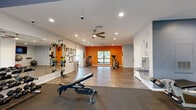 Located at 1602 Hunnington Pl in Hurstbourne Acres, KY, this fitness facility offers a thoughtfully designed single-floor layout that accommodates a full range of workout activities. The space is divided into distinct functional zones, each tailored to specific training needs.

The main exercise area features durable hardwood-style flooring and is accented by a bold orange feature wall that energizes the environment. Recessed lighting and ceiling fans maintain consistent comfort throughout the space. Large floor-to-ceiling windows in the cardio section flood the area with natural light while offering views of the surrounding outdoors, creating an inviting atmosphere for treadmill and cardio machine workouts.

A dedicated strength training section includes a multi-rack dumbbell station, a cable/functional trainer, TRX suspension equipment, and a weight bench. Full-length wall mirrors extend across this zone, enhancing the sense of space and allowing users to monitor their form. Additional cardio equipment, including an elliptical and stationary bike, is positioned in the open hardwood-floored area near the orange accent wall.

A water fountain and hydration station are conveniently accessible. Motivational fitness charts and framed posters are displayed throughout, and a security camera ensures a safe environment. The facility also includes a clearly marked exit corridor, providing easy navigation through the space.