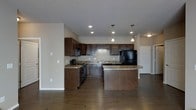 This single-floor space centers around a well-appointed open-concept kitchen that serves as the focal point of the layout. The kitchen is outfitted with rich, dark espresso-toned cabinetry that extends to the upper walls, providing generous storage. Granite countertops with warm undertones complement the cabinetry and add a refined aesthetic. Black appliances, including a refrigerator and range, are seamlessly integrated into the design.

A peninsula island with matching granite extends into the main living area, offering additional prep space and casual seating. Three pendant lights hang above the island, providing focused task lighting and a stylish visual element. Recessed lighting throughout the kitchen ensures the space is well-lit overall.

The flooring consists of dark hardwood-style planks that run continuously through the kitchen and into the adjoining areas, creating a cohesive and grounded look. The neutral wall tones provide a clean backdrop that allows the dark cabinetry and countertops to stand out. A hallway visible in the background suggests additional rooms beyond the open living and kitchen area.