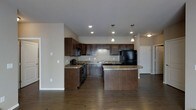This single-floor space centers around a well-appointed open-concept kitchen that serves as the focal point of the layout. The kitchen is outfitted with rich, dark espresso-toned cabinetry that extends to the upper walls, providing generous storage. Granite countertops with warm undertones complement the cabinetry and add a refined aesthetic. Black appliances, including a refrigerator and range, are seamlessly integrated into the design.

A peninsula island with matching granite extends into the main living area, offering additional prep space and casual seating. Three pendant lights hang above the island, providing focused task lighting and a stylish visual element. Recessed lighting throughout the kitchen ensures the space is well-lit overall.

The flooring consists of dark hardwood-style planks that run continuously through the kitchen and into the adjoining areas, creating a cohesive and grounded look. The neutral wall tones provide a clean backdrop that allows the dark cabinetry and countertops to stand out. A hallway visible in the background suggests additional rooms beyond the open living and kitchen area.