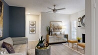This well-appointed single-floor residence showcases a thoughtfully designed open-concept layout that seamlessly connects the living, dining, and kitchen areas. The living room is a standout space, anchored by a white brick fireplace that serves as a striking focal point, complemented by a ceiling fan, light wood-style flooring, and a bold slate-blue accent wall that adds depth and character. Natural light filters in through multiple windows fitted with blinds, enhancing the airy ambiance throughout.

The galley-style kitchen is outfitted with white cabinetry, stone-look countertops, stainless steel appliances including a refrigerator, electric range, microwave, and dishwasher, along with a subway tile backsplash. A pass-through to the living area and a breakfast bar add both function and flow.

The bedroom is a serene retreat featuring plush carpeting, a ceiling fan, and a matching slate-blue accent wall that ties cohesively to the living space. It connects directly to a bathroom with a single vanity, stone countertop, and a large mirror, alongside a well-organized closet. A dedicated workspace area is also integrated into the open living layout, offering flexibility for modern living needs.