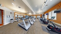 This single-level fitness center is designed to support a comprehensive workout experience, combining cardio and strength training equipment in a spacious, open layout. The interior is immediately distinguished by its bold orange accent walls, which are offset by soft gray-blue sections, creating an energetic yet balanced atmosphere. A decorative heartbeat graphic lines one of the orange walls, reinforcing the fitness-focused character of the space.

The flooring throughout consists of textured, patterned carpet tiles that define workout zones while providing cushioning underfoot. Recessed lighting and suspended fluorescent fixtures illuminate the space evenly, supplemented by ample natural light streaming through multiple large windows fitted with white blinds, offering views of the outdoor surroundings.

The cardio section is prominently equipped with a row of Matrix treadmills, stair climbers, stationary bikes, and a rowing machine, several positioned in front of wall-mounted televisions for entertainment during workouts. Full-length mirrors line multiple walls, visually expanding the space and allowing users to monitor their form.

The strength training area includes adjustable benches, a dumbbell rack, cable machines, and additional resistance equipment. A dedicated section near the entrance features extra free weights and functional training tools. The fitness center is accessed through glass-paneled French doors, and additional rooms appear to be accessible from within for supplementary amenities.