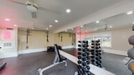 This dedicated fitness center is thoughtfully designed to support a full range of workout activities within a bright, welcoming environment. The space features durable rubber flooring throughout, providing a safe and comfortable surface for both cardio and strength training exercises. Recessed lighting and dual ceiling fans keep the room well-lit and well-ventilated, contributing to a comfortable workout atmosphere.

The strength training area is well-stocked with a tiered dumbbell rack offering a comprehensive range of weights, adjustable weight benches, and a selection of medicine balls with a dedicated storage rack. A wall-mounted TRX suspension training system provides versatility for functional fitness routines, while plyo boxes add options for high-intensity interval training.

A treadmill is positioned along the windowed wall, allowing users to enjoy outdoor views of the surrounding grounds during cardio sessions. Large windows fitted with blinds flood the space with natural light, enhancing the open and energetic feel of the room. Floor-to-ceiling mirrors span one full wall, visually expanding the space and allowing users to monitor their form during workouts.

The fitness center includes double French doors that open directly to the outdoor grounds, providing convenient access and promoting airflow. A motivational neon sign adds a playful, modern touch to the overall aesthetic of the space.