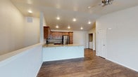 This single-story space showcases an open-concept layout that seamlessly connects the kitchen and living areas. The kitchen is outfitted with warm-toned wooden cabinetry, granite countertops, and stainless steel appliances, including a full-size refrigerator. A peninsula with a built-in sink extends into the living space, offering additional prep area and casual seating potential while reinforcing the open flow between rooms.

Vaulted ceilings add a sense of height and spaciousness throughout the main area, complemented by recessed lighting that provides consistent, even illumination. A ceiling fan in the living area contributes to both comfort and airflow. The rich, dark hardwood-style flooring runs throughout, adding warmth and visual continuity to the space.

Visible in the background is a dedicated laundry area with a washer and dryer, adding functional convenience. A white paneled door leads to an additional room, suggesting further living or storage space. The clean white walls and neutral palette throughout create a bright, versatile environment well-suited for a variety of uses.