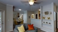 This single-floor residence offers a thoughtfully designed open-concept layout that seamlessly connects the living, dining, and kitchen areas. The living room serves as the heart of the space, featuring a ceiling fan with a light fixture and carpeted flooring that adds warmth underfoot. A comfortable seating area anchors the room, complemented by a built-in shelving unit along the wall — ideal for display and storage. The open layout flows naturally into the dining area, which is well-lit and positioned adjacent to the living space, creating a cohesive and inviting atmosphere for entertaining or everyday living. A half-wall partition with a pass-through counter separates the kitchen, offering both visual openness and functional division. The kitchen is visible beyond the partition, with a glimpse of countertop workspace and cabinetry. Crown molding details add a refined touch throughout the space. Neutral wall tones and consistent ceiling lighting contribute to a clean, unified aesthetic. A central hallway leads to additional areas of the home, including a bathroom visible in the background, hinting at a well-organized floor plan that balances shared and private living spaces.