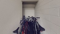 This bike storage facility is a purpose-built space designed to accommodate multiple bicycles in a secure, enclosed environment. The room is constructed with concrete block walls finished in a neutral tone, giving the space a sturdy and utilitarian character. The flooring features a dark, textured surface that provides durability and grip underfoot. A recessed alcove built into one of the walls offers additional capacity for storing bikes compactly, making efficient use of the available space. Signage on the wall indicates stairwell and roof access, confirming the storage area's integration within a larger multi-story building. An emergency exit sign and door are also visible, ensuring compliance with safety standards. Overhead lighting keeps the area well-illuminated. The layout accommodates a variety of bicycle types, from mountain bikes to cruisers, stored side by side along the walls. Overall, the space is straightforward and functional, providing building residents with a convenient and organized solution for bicycle storage.