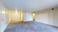 This single-floor space presents an open, functional layout with wall-to-wall carpeting throughout. The main living area is generously proportioned, offering a comfortable sense of space that feels open and versatile. A ceiling fan is mounted to the textured popcorn ceiling, providing both air circulation and ambient lighting to the room.

A set of double closet doors along the back wall provides notable storage capacity, a practical feature of this space. Neutral-toned walls complement the carpeted floors, creating a consistent and understated palette throughout. Two doorways branch off from the main area — one to the left leads to an additional room or hallway, visible with its own lighting, and one to the right opens to another connected space, suggesting a multi-room layout.

The textured ceiling adds a classic architectural detail consistent with the overall design of the space. The layout appears straightforward and accessible, with wide open floor space that offers flexibility for various furniture arrangements or uses. Overall, the space conveys a clean, move-in ready feel with practical features suited for everyday living.