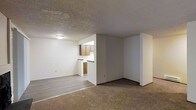 This single-floor unit offers a straightforward, functional layout that connects the living area and kitchen in an open, accessible arrangement. The living space is finished with wall-to-wall carpet, providing a comfortable underfoot feel, while the kitchen and adjoining transition area feature smooth hardwood-style flooring, creating a clear visual distinction between zones. A textured popcorn ceiling spans the entirety of the space, complemented by flush-mounted ceiling lighting that evenly illuminates the interior. The kitchen is partially open to the living area, separated by a low wall that doubles as a countertop pass-through. Warm-toned wood cabinetry lines the kitchen walls, offering practical storage, while the white countertops and walls keep the overall aesthetic clean and neutral. The walls throughout are painted in a light, neutral tone, contributing to a bright and open atmosphere. The layout is straightforward and efficient, making good use of the available floor space. The single-floor design ensures easy accessibility, and the open sightlines between the living and kitchen areas enhance the sense of spaciousness within the unit.