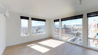 This single-floor space makes a strong impression with its open, light-filled layout and elevated urban setting. The main living area is defined by a wraparound arrangement of large windows fitted with sleek black roller shades, offering panoramic views of a dense cityscape — including rooftops, historic brick buildings, a water tower, and rolling hills in the distance. The corner window configuration maximizes both natural light and sightlines, flooding the interior with sunlight that reflects off the light-toned hardwood flooring throughout. The walls are finished in crisp white, contributing to a clean, minimalist aesthetic that complements the modern design sensibility. Ceilings are smooth and unadorned, reinforcing the sense of openness. The flooring — a light, wide-plank hardwood — adds warmth and continuity across the space. The overall ambiance is airy and contemporary, well-suited for a variety of uses. The elevated position of the unit provides a commanding view of the surrounding urban environment, making the city itself a defining architectural feature of the interior experience.