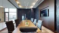 This single-floor conference room offers a refined and professional environment designed to facilitate productive meetings. At its center sits a long, rectangular wooden conference table surrounded by high-backed upholstered chairs with polished chrome bases, providing both comfort and a sleek aesthetic. The room's most striking design element is the deep navy feature wall adorned with a bold, cityscape-inspired wallcovering that adds depth and visual interest to the space.

A large wall-mounted flat-screen display is positioned prominently on the dark wood-paneled wall, ensuring clear visibility for presentations and video conferencing. Overhead, a coffered ceiling with recessed lighting frames an elegant sculptural pendant light fixture, lending a contemporary sophistication to the room. Natural light filters in through floor-to-ceiling windows along one side, offering views of the surrounding cityscape and brightening the overall ambiance. Light-toned flooring contrasts with the darker wall treatments, creating a well-balanced and polished interior. A glass-paneled interior door provides connectivity to adjacent spaces while maintaining visual openness.