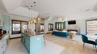 Located at 10480 Auto Mall Pkwy in D'Iberville, Mississippi, this single-floor community clubhouse offers a thoughtfully designed, coastal-inspired interior that blends functionality with stylish décor. The open-plan main area serves as the heart of the space, combining a lounge and social zone with soaring vaulted ceilings, light wood flooring, and an abundance of natural light through large black-framed windows. The lounge features a teal accent wall adorned with decorative oval mirrors, a wall-mounted television, a linear fireplace with botanical wallpaper surround, and woven pendant lighting overhead. A spacious dining nook with large pool-facing windows and elegant teal upholstered chairs provides an inviting gathering area. The kitchen is a standout feature, showcasing sage-green shaker cabinetry, dark quartz countertops, a pink tile-clad island with bar seating, patterned tile flooring, and a cluster of amber glass pendant lights — fully equipped for entertaining. Two private office spaces offer built-in shelving, integrated desk stations with task lighting, and a cozy window bench seat, providing functional workspaces within the broader communal environment. The overall ambiance is welcoming and resort-like, reflecting a cohesive coastal aesthetic throughout.