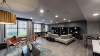 This sophisticated single-floor community lounge is designed with a modern aesthetic that blends comfort and style seamlessly. The space is anchored by light wood-plank flooring that runs throughout, complemented by cool gray walls and recessed lighting that creates a warm, inviting ambiance.

The main lounge area features multiple seating zones, including a large sectional sofa in crisp white, a tufted charcoal sofa, and distinctive leather butterfly chairs. A bold teal and gray abstract area rug grounds one of the primary seating arrangements, while a dramatic black lattice-style shelving unit serves as a striking room divider and decorative focal point.

A dedicated game area is highlighted by a sleek, dark-stained shuffleboard table positioned beneath a brass-toned pendant light, flanked by natural light streaming through large windows dressed with floor-length gray curtains.

The lounge also features a well-appointed coffee bar area, complete with dark cabinetry, a marble-topped counter, and a coffee machine — a nod to everyday convenience. An accent wall adorned with a grid of mixed-media artwork and a woven gold pendant light adds an artistic dimension to the space.

Throughout, carefully chosen décor elements — including a round wall mirror, macramé wall hangings, and sculptural accessories — contribute to a cohesive, upscale residential atmosphere.
