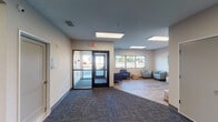 Located at 175 18th St SW in Owatonna, Minnesota, this single-story commercial facility offers a clean, modern, and functional layout designed with comfort and accessibility in mind. The main entrance features a set of glass double doors that flood the entryway with natural light, opening into a spacious reception and transition area finished with durable patterned carpet in muted tones. Large flush-mounted ceiling light panels provide consistent, even illumination throughout the interior.

Just beyond the entry, a welcoming lounge area is appointed with contemporary blue upholstered armchairs, a light teal accent chair, a round wooden side table, and a woven pouf ottoman, creating an inviting space for visitors. Colorful abstract artwork adorns the walls, adding vibrant character to the otherwise neutral palette of soft whites and warm beiges.

The open interior extends into a broader multi-purpose area supported by structural columns and connected by wide corridors, allowing for flexible use of the space. Transitions between carpet and wood-look luxury vinyl flooring delineate distinct functional zones. Windows along the exterior wall offer views of the surrounding grounds, further enhancing the bright and airy atmosphere. The overall design reflects a professional yet approachable environment suited for community or client-facing operations.