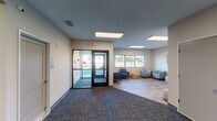 Located at 175 18th St SW in Owatonna, Minnesota, this single-story commercial facility offers a clean, modern, and functional layout designed with comfort and accessibility in mind. The main entrance features a set of glass double doors that flood the entryway with natural light, opening into a spacious reception and transition area finished with durable patterned carpet in muted tones. Large flush-mounted ceiling light panels provide consistent, even illumination throughout the interior.

Just beyond the entry, a welcoming lounge area is appointed with contemporary blue upholstered armchairs, a light teal accent chair, a round wooden side table, and a woven pouf ottoman, creating an inviting space for visitors. Colorful abstract artwork adorns the walls, adding vibrant character to the otherwise neutral palette of soft whites and warm beiges.

The open interior extends into a broader multi-purpose area supported by structural columns and connected by wide corridors, allowing for flexible use of the space. Transitions between carpet and wood-look luxury vinyl flooring delineate distinct functional zones. Windows along the exterior wall offer views of the surrounding grounds, further enhancing the bright and airy atmosphere. The overall design reflects a professional yet approachable environment suited for community or client-facing operations.