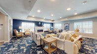 This single-floor community space is thoughtfully designed with a cohesive coastal aesthetic, blending comfort and functionality throughout its distinct areas. The expansive lounge area serves as the heart of the space, featuring a bold navy accent wall, a sleek wall-mounted fireplace, and plush seating arrangements including striped armchairs, a large sectional sofa, and a blue loveseat. A striking navy-and-cream abstract patterned carpet anchors the lounge, complemented by natural wood accent tables and carefully curated nautical décor. Recessed lighting and tray ceilings add architectural dimension, while an abundance of windows dressed with white plantation shutters floods the room with natural light and outdoor views. Adjacent to the lounge, the dining area transitions seamlessly onto warm hardwood-style flooring, offering multiple dining tables and chairs arranged to accommodate a variety of group sizes. Coastal-inspired artwork adorns the walls throughout. The fully equipped kitchen features dark cabinetry, light countertops, stainless steel appliances including a refrigerator and dishwasher, and a central island that provides ample prep and serving space. A set of glass-paneled doors in the kitchen area connects to the exterior, enhancing the sense of openness and accessibility across this well-appointed communal facility.