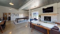 Located at 3425 W Leigh St in Richmond, Virginia, this single-floor community amenity space combines industrial-modern design with a warm, inviting atmosphere. The expansive open-plan lounge serves as the heart of the space, showcasing soaring ceilings with a distinctive teal-painted corrugated metal finish, exposed structural beams, and polished concrete floors throughout. A striking stone-surround fireplace anchors one end of the lounge, complemented by a wall-mounted flat-screen TV, rich leather sofas, and vibrant blue accent chairs. Large floor-to-ceiling glass doors open to an outdoor area, flooding the interior with natural light and connecting seamlessly to the exterior. A dedicated dining zone features a solid wood communal table, spindle-back chairs, and high-top seating beneath clerestory windows that frame views of the surrounding neighborhood. For entertainment, a full-size pool table adds a recreational focal point. The kitchenette is outfitted with white shaker cabinetry, dark granite countertops, stainless steel appliances including a full-size refrigerator and microwave, and colorful artwork that brightens the space. The fitness center is well-equipped with cardio machines, a multi-station cable machine, free weights, and mirrored walls, all set on durable rubber flooring with ample natural light.