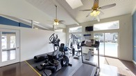 This fitness center offers a bright and inviting environment for exercise, characterized by soaring vaulted ceilings with a white beadboard finish that lend a sense of openness to the space. Two ceiling fans with light fixtures are suspended from the pitched ceiling, ensuring comfortable air circulation throughout workouts. Clerestory windows positioned along the upper walls allow generous natural light to flood the interior, creating an energizing atmosphere.

The facility is equipped with a well-rounded selection of cardio machines, including treadmills, an elliptical trainer, and a stationary bike, all arranged to maximize usability within the space. A multi-station strength training machine is also available for resistance-based workouts. A wall-mounted flat-screen television provides entertainment during exercise sessions, and an informational board is posted nearby for member reference.

Large glass sliding doors along one wall open toward an outdoor pool area, offering refreshing views of the surrounding landscape and enhancing the connection between indoor and outdoor amenities. The overall ambiance is clean, functional, and well-lit, making this fitness center a practical and welcoming facility.
