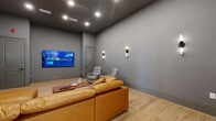 This thoughtfully designed media lounge offers a dedicated entertainment experience within a compact yet well-appointed single-room layout. The space is finished with cool-toned grey walls and a dark ceiling that enhance the cinematic atmosphere, while light hardwood flooring provides a warm contrast underfoot.

At the heart of the room, a large wall-mounted smart TV serves as the focal point, positioned for optimal viewing from multiple seating areas. A generous caramel-toned leather sectional sofa anchors the space, complemented by a low-profile wooden coffee table with drawer storage. Two upholstered grey recliner chairs offer additional seating and are elegantly lit by a trio of decorative wall sconces along the side wall.

Recessed ceiling lights provide ambient overhead illumination, while the stylish sconce fixtures add a refined, layered lighting scheme throughout. A built-in shelving nook with display space and a small credenza are visible near the rear, adding functional storage. French doors at the back of the room allow access to an adjacent bright space, enhancing the sense of openness. A secondary interior door provides a private entry point.

The overall ambiance is modern and inviting, combining sleek design with comfort-focused furnishings to create an ideal space for media viewing and relaxed lounging.