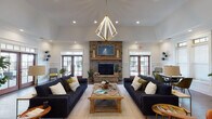 Located at 145 Retreat Dr in Pittsboro, NC, this single-story clubhouse-style space offers a thoughtfully designed open floor plan that seamlessly blends comfort and functionality. The centerpiece of the main lounge is a striking floor-to-ceiling stone fireplace, anchoring the room beneath soaring vaulted ceilings adorned with a geometric gold chandelier. Two large dark sofas flank a rustic wooden coffee table, creating an inviting gathering space. Natural light pours in through multiple sets of French doors with transom windows, offering views of the surrounding outdoor areas. The warm hardwood flooring flows throughout, connecting the various zones of the space. A secondary lounge area provides additional seating with a mix of mid-century modern armchairs in rich leather and velvet tones, alongside a wall-mounted television and a sleek credenza. The fully appointed kitchen features crisp white shaker cabinetry, dark countertops, subway tile backsplash in a herringbone pattern, stainless steel appliances, and a central island with a brass fixture sink. Wainscoting detail along the lower walls adds a refined architectural touch throughout, while recessed lighting and abundant natural light contribute to a bright, welcoming ambiance throughout the entire space.