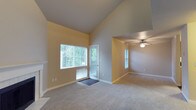 This two-floor space offers a welcoming and well-lit interior defined by thoughtful design details and neutral finishes. The main living area immediately impresses with its dramatic vaulted ceiling, which amplifies the sense of openness and height. Large windows with blinds invite generous amounts of natural light, offering pleasant views of lush greenery outside. A glass-paneled door provides additional light and convenient access to the exterior. The carpeted flooring in soft, neutral tones extends throughout the living spaces, contributing to a warm and comfortable ambiance. An adjacent room, visible through an open arched passageway, features standard ceiling height and is equipped with a ceiling fan with integrated lighting, providing both comfort and functionality. The warm yellow-toned walls throughout lend a cohesive and inviting character to the space. White baseboards provide a clean contrast against the soft wall colors. The open layout between the main and adjacent rooms allows for flexible use of the space and promotes easy flow between areas. Overall, this two-floor property combines airy architectural elements with practical features, making it a comfortable and versatile living environment filled with natural light and a sense of spaciousness.