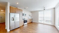 This contemporary single-floor residence offers a thoughtfully designed layout with clean lines and a neutral color palette throughout. The open-concept kitchen is a standout feature, outfitted with a full suite of stainless steel appliances including a side-by-side refrigerator with ice and water dispenser, electric range, over-the-range microwave, and dishwasher. Cabinetry combines warm wood-tone lower cabinets with crisp white upper cabinets, complemented by light countertops and a simple single-basin sink. Warm hardwood-style flooring flows continuously through the kitchen, living area, hallways, and bedrooms, providing a cohesive and inviting feel. The living area benefits from abundant natural light through large windows fitted with horizontal blinds, and a ceiling fan with a gold-toned fixture adds both function and style. The bedrooms are generously sized with windows offering outdoor views, ceiling fans, and ample natural light. A spacious walk-in closet features wire shelving and hanging rods along multiple walls. The bathrooms are finished with wood-tone vanities, white quartz-style countertops, large framed mirrors, and brushed nickel fixtures. One bathroom includes a combination tub and shower with a rain showerhead, while the other features a wide vanity with drawers. A central hallway connects all areas with recessed lighting overhead.