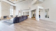 Located at 8900 Independence Pkwy in Plano, TX, this single-floor community amenity space offers a well-rounded collection of functional and elegantly designed rooms. The expansive living and lounge area immediately impresses with soaring ceilings, white-painted structural columns, and light wood-look flooring that flows throughout. Large windows invite abundant natural light, and the open layout accommodates generous seating arrangements, including a curved sectional sofa and accent chairs, creating an inviting social atmosphere. The dining room features a bold, live-edge wood table surrounded by leather chairs beneath striking black-and-gold pendant lighting. Built-in cabinetry and multiple mounted flat-screen TVs make this space equally suited for casual gatherings or structured meetings. The kitchen is outfitted with stainless steel appliances, including a French door refrigerator, white subway tile backsplash, open wood shelving, and a commercial-style coffee station — ideal for community use. The fitness center stands out with dramatic arched windows, vaulted ceilings, ceiling fans, and an extensive array of cardio machines, free weights, and strength equipment, all flooded with outdoor views. A secondary exercise area with mirrored walls and built-in shelving provides additional workout space. Throughout, the design maintains a clean, modern aesthetic with a neutral white palette.