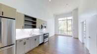 This well-appointed 1-bedroom, 1-bathroom residence offers a thoughtfully designed open-concept layout that seamlessly blends the kitchen and living areas into a bright, cohesive space. The kitchen is outfitted with modern gray cabinetry featuring open shelving, sleek white quartz countertops, a clean subway tile backsplash, and a full suite of stainless steel appliances including a gas range, dishwasher, and built-in microwave. Recessed lighting throughout provides consistent, functional illumination. The living area is anchored by a striking floor-to-ceiling window that floods the space with natural light and frames a verdant, tree-lined view, creating a seamless connection between the interior and the outdoors. Warm hardwood-style flooring runs throughout, adding richness and continuity to the open floor plan. Soaring ceilings enhance the sense of volume and airiness, making the space feel expansive. The clean, minimalist aesthetic is carried through every detail, from the crisp white walls to the contemporary hardware, resulting in a residence that is both functional and visually refined.