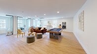 This 730 square foot single-floor space in Barrington, NJ, showcases a versatile and contemporary design. The open floor plan maximizes the area's functionality, seamlessly blending work and leisure zones. Light wood flooring runs throughout, complementing the crisp white walls and creating a bright, airy atmosphere.

The space is thoughtfully divided into distinct areas. A sleek work station with a glass partition offers a dedicated office space, while a lounge area features a comfortable brown sofa and circular ottomans. A centerpiece pool table adds a recreational element, perfect for unwinding or entertaining.

Large windows flood the room with natural light, offering views of the outdoors. The minimalist decor includes carefully chosen artwork and decorative items, maintaining a clean, uncluttered look. Modern amenities such as recessed lighting and a ceiling-mounted HVAC system ensure comfort and functionality.

Storage is cleverly integrated with a long, wooden credenza that provides ample space for organization while serving as a stylish design element. The overall aesthetic is one of contemporary elegance, balancing form and function in a compact yet open environment.