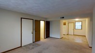 This single-floor unit presents an open, functional layout designed for comfortable everyday living. The main living area is finished with neutral wall-to-wall carpeting that extends throughout, creating a cohesive and understated aesthetic. The entry area transitions from tile flooring into the carpeted space, offering a practical delineation between the entryway and the living zone.

Several interior doors are visible from the main living area, including what appears to be a closet with built-in shelving, and an additional door leading to a secondary space. The walls are painted in a neutral gray-white tone, contributing to a clean, understated atmosphere throughout.

A hallway at the far end of the living area connects to the kitchen, where a white refrigerator is visible, suggesting a functional galley-style or compact kitchen setup with warm wood-toned cabinetry. Ceiling vents indicate a central HVAC system. The textured ceiling and recessed vent placement are consistent throughout the space. Overall, the unit offers a straightforward, practical layout with clear sightlines between the main living area and kitchen.