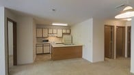 This single-floor residential space offers a practical and functional layout centered around an open-concept kitchen and living area. The kitchen is well-appointed with an abundance of two-toned cabinetry — cream upper cabinets with warm brown lower cabinets — providing generous storage space. It comes equipped with a white refrigerator, electric range, built-in microwave, and a double sink integrated into the peninsula counter. The peninsula serves as a natural divider between the kitchen and the adjoining living area, creating a seamless flow between the two spaces while maintaining an open and airy feel. The living area is finished with plush neutral-toned carpeting, contributing to a comfortable and understated ambiance. The kitchen features tile flooring that transitions cleanly into the carpeted living space. A textured ceiling runs throughout, and recessed ventilation ensures good air circulation. The walls are painted in a light, neutral tone, enhancing the brightness of the space. The layout is efficient and well-organized, making the most of the available square footage while keeping the overall atmosphere clean and welcoming. This space is well-suited for everyday living with its cohesive design and thoughtful use of space.