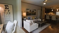 This single-story space showcases a thoughtfully designed open-concept living area that blends comfort and contemporary style. The living room serves as the focal point, featuring light-colored upholstered seating, including a sofa accented with decorative pillows in soft blues and yellows, and a matching armchair arranged around a rustic wood coffee table. A textured area rug anchors the seating arrangement atop warm hardwood-style flooring, which transitions to tile in adjoining areas. Neutral taupe walls provide a calm backdrop, enhanced by a pair of large landscape-themed canvas artworks that add a natural, earthy element to the space. A ceiling fan with integrated lighting contributes both function and a finishing touch overhead. A structural column adds architectural interest while subtly defining the living area. Natural and ambient lighting work together to create a warm, inviting atmosphere throughout. Visible in the background are additional rooms, including what appears to be a kitchen with dark cabinetry, suggesting a well-connected, flowing floor plan. The overall ambiance is relaxed yet refined, suited for comfortable everyday living.
