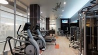 This modern fitness center is designed to support a full range of workout routines in a well-organized, single-level layout. The cardio zone features a row of Matrix elliptical trainers and treadmills, providing ample equipment for members during peak hours. Opposite the cardio area, a dedicated strength training section includes cable machines and various resistance equipment arranged for efficient use of the floor space. The interior design blends function with contemporary style, showcasing dark accent columns, neutral-toned carpeting with bold orange geometric accents, and large floor-to-ceiling windows along one wall that allow natural light to fill the space. Recessed ceiling lighting is complemented by ceiling fans, ensuring the environment remains comfortable during intense workouts. A wall-mounted flat-screen television is positioned prominently for viewing during cardio sessions. The open floor plan offers clear sightlines throughout the facility, contributing to a safe and accessible workout environment. The overall ambiance is clean, modern, and purposefully designed to motivate and accommodate a variety of fitness goals.