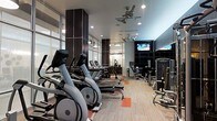 This modern fitness center is designed to support a full range of workout routines in a well-organized, single-level layout. The cardio zone features a row of Matrix elliptical trainers and treadmills, providing ample equipment for members during peak hours. Opposite the cardio area, a dedicated strength training section includes cable machines and various resistance equipment arranged for efficient use of the floor space. The interior design blends function with contemporary style, showcasing dark accent columns, neutral-toned carpeting with bold orange geometric accents, and large floor-to-ceiling windows along one wall that allow natural light to fill the space. Recessed ceiling lighting is complemented by ceiling fans, ensuring the environment remains comfortable during intense workouts. A wall-mounted flat-screen television is positioned prominently for viewing during cardio sessions. The open floor plan offers clear sightlines throughout the facility, contributing to a safe and accessible workout environment. The overall ambiance is clean, modern, and purposefully designed to motivate and accommodate a variety of fitness goals.