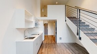 Located at 2211 SW Fourth Ave in Portland, Oregon, this efficiently designed loft-style unit makes smart use of its compact footprint through thoughtful vertical space planning. The main level features an open-plan kitchen outfitted with white cabinetry, open wall shelving, a stainless steel sink, a built-in cooktop, and a microwave. Warm wood-look flooring runs throughout the main level, complementing the crisp white walls and recessed lighting. A full bathroom on the main floor is clean and functional, featuring wood-look flooring, a bathtub, toilet, and grab bars. A striking staircase with a sleek metal cable railing serves as a central architectural feature, leading to an upper loft level. The loft area is finished with plush carpeting and bright white walls, offering a flexible space bathed in natural light from large windows that frame views of the surrounding Portland cityscape and greenery. Pendant lighting and a ceiling fan add both style and comfort to the upper level. The overall ambiance is modern and minimalist, with a light, airy palette and clean lines throughout that maximize the sense of space in this efficiently laid-out urban dwelling.