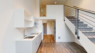 Located at 2211 SW Fourth Ave in Portland, Oregon, this efficiently designed loft-style unit makes smart use of its compact footprint through thoughtful vertical space planning. The main level features an open-plan kitchen outfitted with white cabinetry, open wall shelving, a stainless steel sink, a built-in cooktop, and a microwave. Warm wood-look flooring runs throughout the main level, complementing the crisp white walls and recessed lighting. A full bathroom on the main floor is clean and functional, featuring wood-look flooring, a bathtub, toilet, and grab bars. A striking staircase with a sleek metal cable railing serves as a central architectural feature, leading to an upper loft level. The loft area is finished with plush carpeting and bright white walls, offering a flexible space bathed in natural light from large windows that frame views of the surrounding Portland cityscape and greenery. Pendant lighting and a ceiling fan add both style and comfort to the upper level. The overall ambiance is modern and minimalist, with a light, airy palette and clean lines throughout that maximize the sense of space in this efficiently laid-out urban dwelling.