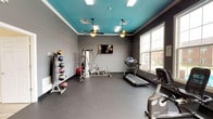 This fitness center in Vineland is a well-equipped, single-floor space designed to support a comprehensive workout routine. The room's most distinctive architectural feature is its bold teal ceiling, which contrasts sharply with the neutral gray walls and dark rubberized flooring, creating a modern and energizing atmosphere. Two ceiling fans provide consistent air circulation throughout the space, complementing the flush-mounted lighting panels that ensure bright, even illumination.

Natural light enters through multiple large windows along the perimeter walls, offering views of the surrounding outdoor environment and enhancing the open feel of the room. The generous floor plan allows ample space between equipment, supporting comfortable movement and safe exercise.

The fitness center is stocked with a diverse range of equipment, including a multi-station cable and weight machine, a treadmill, stationary bike, elliptical trainers, and a stair climber. A wall-mounted medicine ball rack provides options for functional training, while a flat-screen television mounted on the wall adds an entertainment element during workouts. A paper towel and sanitizer station is conveniently positioned near the exit, promoting cleanliness and hygiene throughout the facility.