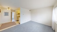 This single-floor unit presents a clean, functional layout designed for everyday living. The living area is generously sized, featuring neutral-toned walls and grey carpeting that extends throughout, creating a cohesive and understated aesthetic. A window with horizontal blinds allows natural light to filter in, brightening the open space. The kitchen is laid out in a galley-style configuration, with white cabinetry offering ample storage above and below the countertops. It is equipped with a gas range, a white refrigerator, and a double-basin sink, all set against light-toned countertops and easy-care flooring. The kitchen opens directly toward the main living space, providing a connected feel between the two areas. The bedroom is a well-proportioned room featuring the same grey carpeting found throughout the unit. A large window dressed with vertical blinds provides natural light and an outdoor view, while a second angle reveals an adjoining doorway leading to the bathroom. The bathroom includes a full tub and shower combo with tiled surround and a decorative curtain, complemented by tile flooring, a toilet, and a vanity. Overall, the unit maintains a simple, practical ambiance with a neutral color palette throughout.