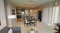 This stylish single-floor residence showcases a thoughtfully designed open-concept floor plan that effortlessly blends the kitchen, dining, and living spaces into one cohesive and inviting environment. The kitchen is outfitted with dark cabinetry, stainless steel appliances, and a mosaic tile backsplash that adds a striking visual accent. A central island with pendant lighting serves as both a functional workspace and a natural gathering point. The dining area is positioned at the heart of the space, illuminated by a sleek track lighting fixture overhead, and comfortably accommodates a round dining table with upholstered chairs. Rich hardwood-style flooring in the kitchen and dining zone transitions smoothly into plush carpeting in the living area, creating a warm and comfortable atmosphere. The living space is open and airy, benefiting from the expansive layout and neutral wall tones that enhance the sense of brightness. A dedicated laundry area is conveniently tucked in the background, adding practical functionality. Additional storage and closet space are visible, further contributing to the unit's livability. Overall, the space strikes a clean, modern aesthetic with a welcoming ambiance suited for everyday living and entertaining alike.