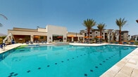 Located at 7430 N Silverbell Rd in Marana, Arizona, this outdoor amenity space showcases a resort-style pool area designed for leisure and relaxation in the Sonoran Desert. The centerpiece is a generously sized swimming pool with striking blue-tiled accents, complemented by a separate elevated spa and a dedicated water feature wall, offering multiple aquatic experiences within a single cohesive environment. The pool deck is finished in light-toned natural stone pavers, providing a clean, upscale aesthetic while remaining cool underfoot in the Arizona sun. Tall, mature palm trees are thoughtfully distributed throughout the space, lending a tropical ambiance and partial shade to the surrounding lounge areas. Vibrant orange market umbrellas and blue chaise lounges dot the expansive deck, creating comfortable, color-coordinated gathering zones. The amenity building adjacent to the pool features large glass storefronts and a modern, tile-clad exterior that integrates seamlessly with the overall desert-contemporary architectural style of the multi-story residential buildings framing the courtyard. An ADA-accessible pool lift is also present, ensuring inclusive access. In the background, the rugged silhouette of the Tucson Mountain range provides a dramatic natural backdrop beneath open Arizona skies, enhancing the overall outdoor experience.