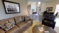 This single-story clubhouse offers a thoughtfully designed communal space that balances comfort and style. The lounge area serves as the heart of the space, featuring a generously sized tan leather sofa accented with decorative pillows, a round wooden coffee table, and a plush patterned area rug that anchors the seating arrangement. A sleek black leather chair adds additional seating nearby, making the space well-suited for both casual conversation and quiet relaxation.

The interior benefits from impressively high ceilings that lend an open, airy feel throughout. A sculptural multi-bulb floor lamp provides warm ambient lighting, complementing the natural light that filters in through the large entrance doors at the far end of the room. Wall sconces near the entryway add decorative character, while a framed landscape artwork on the wall contributes to the refined yet approachable aesthetic.

Polished concrete flooring runs throughout, offering a modern, durable surface that ties the space together seamlessly. In the background, a reception or service counter area is visible, suggesting additional functionality within the clubhouse. The overall ambiance is welcoming, polished, and well-appointed.