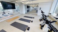 This contemporary fitness facility is thoughtfully designed to accommodate a variety of exercise disciplines within a clean, well-lit environment. The space is divided into distinct workout zones, each purpose-built for specific activities. The yoga and stretching area features a cushioned floor with exercise mats and foam blocks arranged for individual sessions, alongside a wall-mounted flat-screen display — ideal for guided classes or instructional content. Floor-to-ceiling mirrors line the walls, enhancing the sense of openness and allowing participants to monitor their form. Adjacent to the mat area, a selection of stationary cycling machines and cardio equipment is positioned for convenient access. Separated by glass partition walls, the main gym zone is visible beyond, housing a range of strength training machines and cable equipment arranged to maximize usable floor space. The facility benefits from bright overhead fluorescent lighting integrated into an exposed ceiling grid, contributing to an energetic yet functional atmosphere. White walls and light-toned flooring throughout maintain a modern, cohesive aesthetic. The glass partitions between zones create visual connectivity while allowing each area to serve its intended purpose without interference.