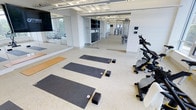 This contemporary fitness facility is thoughtfully designed to accommodate a variety of exercise disciplines within a clean, well-lit environment. The space is divided into distinct workout zones, each purpose-built for specific activities. The yoga and stretching area features a cushioned floor with exercise mats and foam blocks arranged for individual sessions, alongside a wall-mounted flat-screen display — ideal for guided classes or instructional content. Floor-to-ceiling mirrors line the walls, enhancing the sense of openness and allowing participants to monitor their form. Adjacent to the mat area, a selection of stationary cycling machines and cardio equipment is positioned for convenient access. Separated by glass partition walls, the main gym zone is visible beyond, housing a range of strength training machines and cable equipment arranged to maximize usable floor space. The facility benefits from bright overhead fluorescent lighting integrated into an exposed ceiling grid, contributing to an energetic yet functional atmosphere. White walls and light-toned flooring throughout maintain a modern, cohesive aesthetic. The glass partitions between zones create visual connectivity while allowing each area to serve its intended purpose without interference.