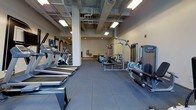 This fitness center offers a comprehensive workout environment designed to accommodate a wide range of exercise needs. The space is defined by its industrial aesthetic, with exposed ceiling infrastructure including visible ductwork, pipes, and structural concrete elements that lend a raw, urban character to the room. Durable rubber flooring tiles cover the entire workout area, providing both safety and practicality underfoot.

Along one side of the space, a row of treadmills and upright cardio machines — including stationary bikes — lines the wall, accompanied by a large flat-screen television mounted above bold motivational lettering. The opposite side of the floor houses a variety of strength and resistance training equipment, including a multi-station cable machine, rowing machines, and a functional trainer. Stability balls and additional accessories are also available for functional fitness and core training.

The space is illuminated by overhead fluorescent lighting that ensures the entire area is brightly and evenly lit. Structural columns are integrated throughout the layout, adding to the industrial character while defining distinct zones within the fitness floor. The overall ambiance is focused and utilitarian, creating an environment well-suited for serious and consistent training.