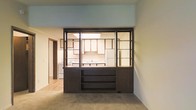 This single-floor space is defined by a standout architectural feature: a bold, dark-framed glass partition that separates the living area from the kitchen. This room divider combines open shelving, glass panes, and a solid lower cabinet section, creating a semi-open design that allows light and sightlines to pass between spaces while maintaining a sense of separation. The lower portion of the divider includes built-in shelving, adding functional storage directly accessible from the living side.

The kitchen, visible through the glass partition, features white upper cabinetry, a white refrigerator, and a fluorescent ceiling light that illuminates the workspace. A sink is positioned along the kitchen counter, and additional appliances are visible along the back wall.

The living area is finished with neutral carpet flooring, while the kitchen transitions to hard flooring, offering a clear material distinction between the two zones. Light-colored walls throughout keep the interior feeling open. A hallway is visible to the left, suggesting additional rooms beyond the main living and kitchen areas. The overall aesthetic reflects a retro, mid-century-influenced design sensibility.