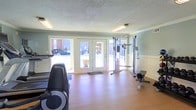 This single-story fitness room offers a clean, functional environment for exercise. The space features light hardwood-style flooring and soft sage-green walls accented by white wainscoting, creating a bright and refreshing atmosphere. A textured ceiling with fluorescent lighting ensures consistent illumination throughout. Large French doors along one wall allow abundant natural light to flow in while providing views of the outdoor surroundings, enhancing the open feel of the room. The fitness area is equipped with a cable resistance machine suitable for a variety of strength training exercises, a rowing machine, and a cardio machine, offering a well-rounded workout experience. A branded floor mat near the entrance adds a polished touch. The layout provides ample space to move between equipment comfortably, making the room practical for individual use. The overall ambiance is clean and inviting, with the combination of natural light, neutral tones, and organized equipment contributing to a motivating workout environment.