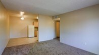 This single-floor residential unit offers a straightforward, open layout designed for comfortable everyday living. The main living area features wall-to-wall carpeting and a textured ceiling, creating a warm and functional space. A ceiling fan is positioned near the transition between the living area and the kitchen, providing air circulation throughout the open-concept floor plan. The kitchen is efficiently arranged with light wood cabinetry, a white refrigerator, and a dishwasher, offering essential appliances within a compact and practical footprint. A visible HVAC vent in the ceiling ensures climate control throughout the unit. Neutral, warm-toned walls run throughout the space, lending a clean and cohesive aesthetic. A doorway visible from the main living area leads to additional rooms, suggesting a connected and accessible interior layout. The overall ambiance is simple and functional, with an emphasis on open sightlines and efficient use of space. Natural light filters into the unit, enhancing the brightness of the neutral interior palette. This unit is well-suited for those seeking a no-frills, move-in-ready living environment with essential amenities conveniently integrated into the layout.