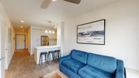 This thoughtfully designed single-floor residence offers a clean, contemporary aesthetic defined by neutral tones, wood-style flooring, and an open layout that encourages comfortable everyday living. The open-concept living and kitchen area serves as the heart of the home, where a well-appointed kitchen features white shaker-style cabinetry, stainless steel appliances including a refrigerator, range, microwave, and dishwasher, and a white quartz peninsula with bar seating illuminated by pendant lighting. The adjoining living area provides a relaxed setting complemented by a ceiling fan overhead. Two bedrooms offer restful retreats, with the primary bedroom distinguished by large floor-to-ceiling windows that frame outdoor views and invite abundant natural light. The second bedroom maintains the same calm, neutral palette. Two full bathrooms serve the residence, each finished with white vanities, ample mirror space, and shower-tub combinations. Wood-style flooring runs consistently throughout the unit, contributing to a cohesive and polished overall ambiance.