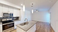 Located in Austin, TX, this thoughtfully designed 2-bedroom, 2-bathroom residence offers a clean, modern aesthetic across a well-organized single-floor layout. The open-concept kitchen and living area serves as the heart of the home, anchored by a sleek center island with a stainless steel sink and quartz-style countertops. The kitchen is fully equipped with stainless steel appliances—including a refrigerator, electric range, and microwave—complemented by crisp white cabinetry extending to the ceiling and a striking dark tile backsplash. Pendant lighting above the island adds a contemporary touch. The living area extends naturally from the kitchen, bathed in natural light from large windows and finished with warm-toned wood flooring that runs throughout the entire space. A ceiling fan provides added comfort. Both bedrooms are bright and well-proportioned, with natural light filtering through slatted blinds. The primary bedroom includes access to a dedicated walk-in closet with built-in shelving. The bathrooms feature white vanities, mirrors with overhead lighting, and matching wood-look flooring. Clean white walls, recessed lighting, and a consistent modern palette give the entire residence a fresh, move-in-ready feel.